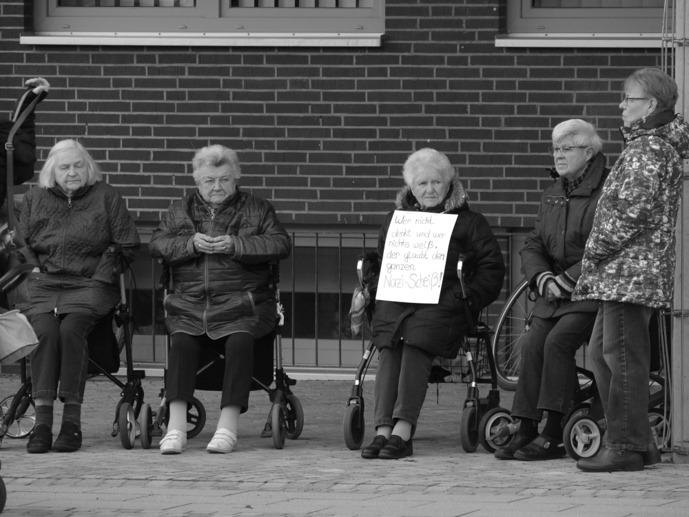Das s/w-Foto stammt von einer Demo für Demokratie und gegen Rechtsextremismus. Man sieht eine Gruppe von fünf älteren Damen, vier sitzen auf ihren Rollatoren und eine trägt ein Schild "er nicht denkt und wer nichts weiß, der gleubt den ganzen Naz-Scheiß!".