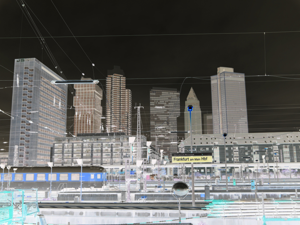 Das Bild ist eine künstlerisch durch eine Farbinvertierung bearbeitets Landschaftsbild aufgenommen vom Frankfurter Hauptbahnhof. Im Hintergrund sind 7 Hochhäuser ersichtlich, davor "normale" Häuser und im Fordergrund der die Bahnsteige des Braunschweiger Hauptbahnhofs. Man kann ein Bahnhofsschild erkennen und einen Personenwagen.