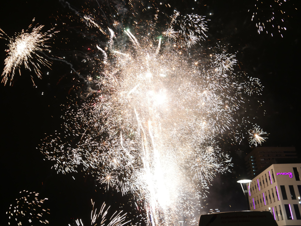 Das Foto zeigt sehr viele helle, weiße Feuerwerksexplosionen am Nachthimmel. Rechts das Hotel Moxy.
