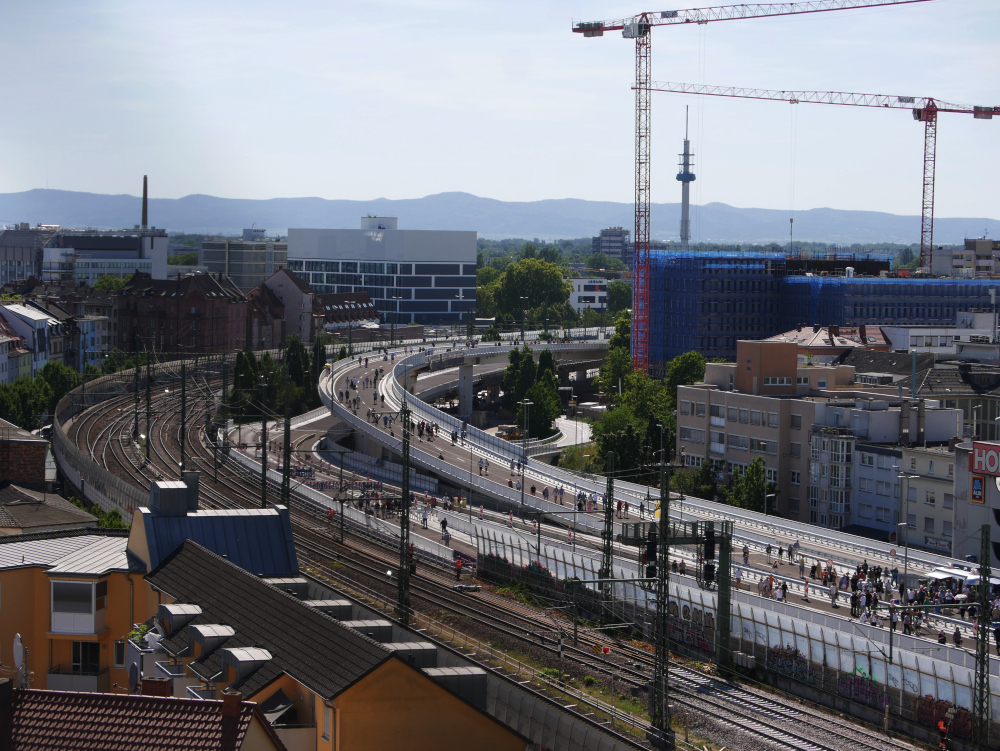 Das Foto zeigt zeigt die Hochstraße Süd in Ludwigshafen am Tag der Eröffnung. Man sieht die vielen Fußgänger*innen, welche an diesem Tag auf die Hochstraße durften und ein paar Imbißwagen. Neben der Hochstraße ist auch die Eisenbahntrasse und die Stadtbebauung erkennbar. Rechts entsteht auch ein weiterer Hochhauskomplex, an dem stehen gerade 2 Baukräne.