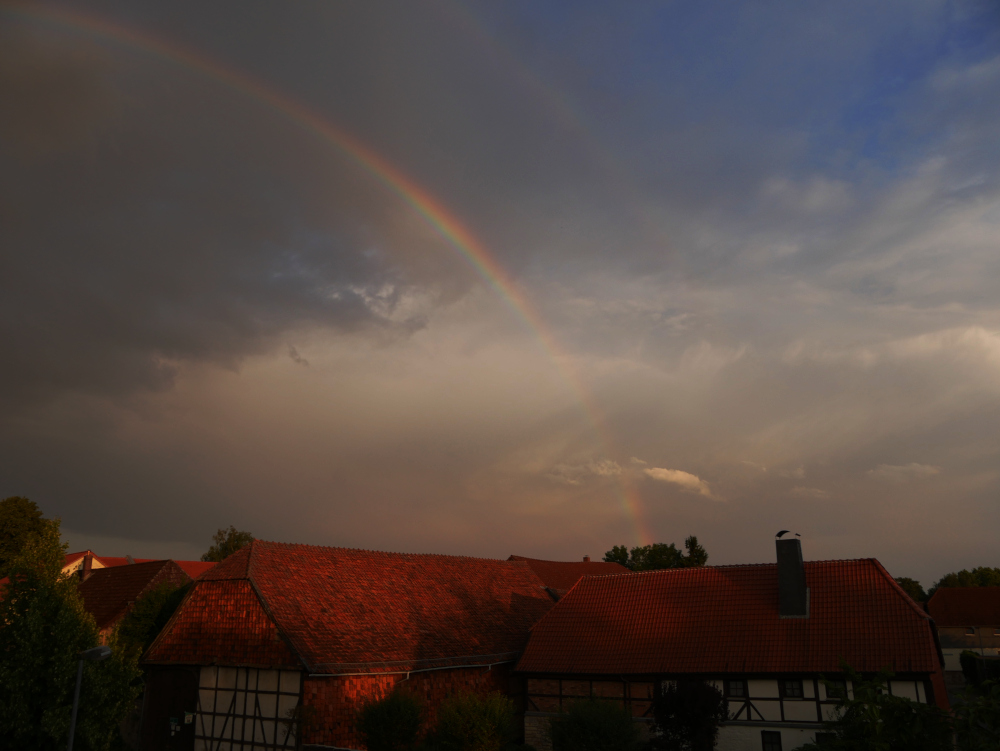 Die Landschaftsaufnahme eigt einen Regenbogen am Regen-Himmel über einem Dorf. Unten sieht man Fachwrk-Gebäude von Höfen, darüber der Regenbogen, der am wolkigen Himmel steht.