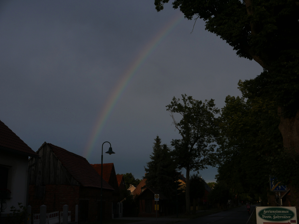 Das Foto zeigt einen Regenbogen am dunkelen, wolkenverhangenen Himmel. Man sieht unten die Häuser und der Straße von Schlepzig, Links eine Bushaltestelle und rechts ein Wegweiser "Fremdenzimemr, Boote, Fahrräder"