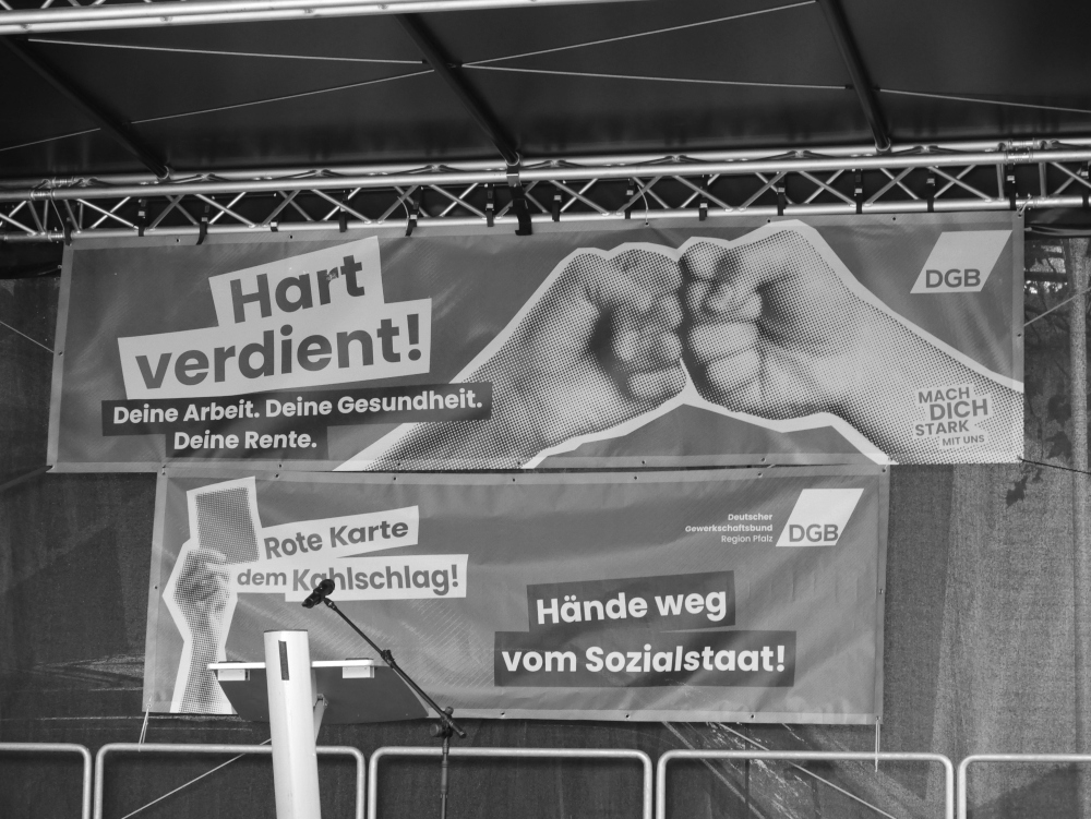 Das s/w-Foto zeigt die leere Bühne der Demo. Man sieht zwei Transparente "Hart verdient! Deine Arbeit. Deine Gesundheit. Deine Rente" und darunter "Rote Karte dem Kahlschlag! Hände weg vom Sozialstaat!
