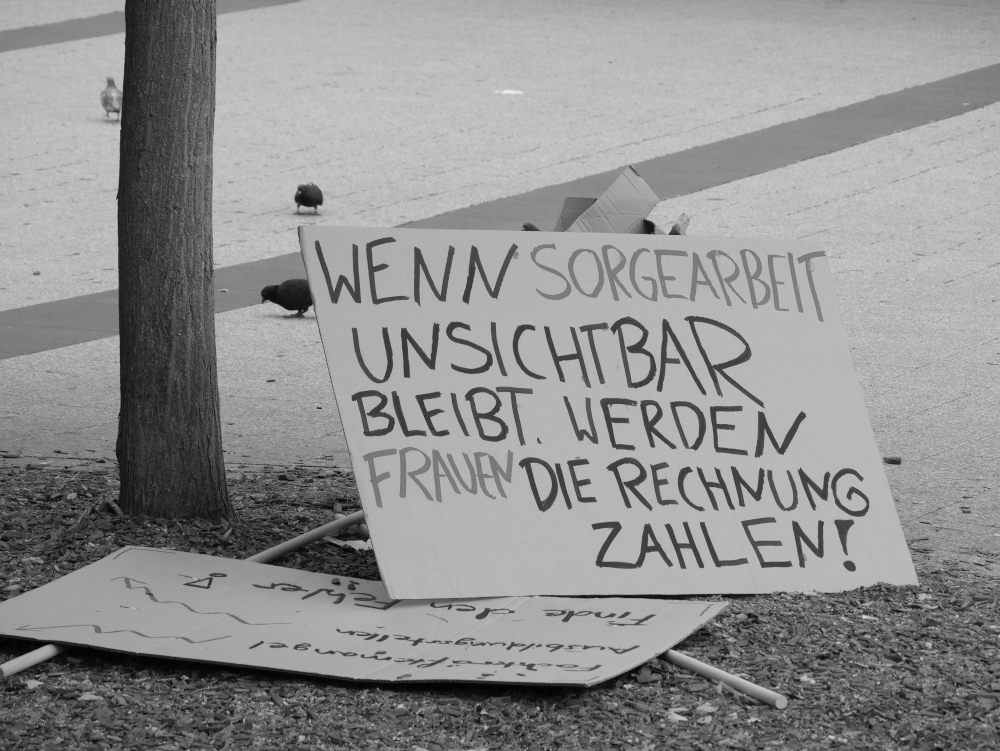 Das s/w-Foto zeigt zwei auf dem boden liegende Plakate. Auf dem einen ist die Aufschrift "Wenn Sorgearbeit unsichtbar bleibt werden Frauen die Rechnung zahlen!", beim anderen ist die Aufschrift nicht lesbar. Im Hintergrund picken drei Tauben auf dem Platz.