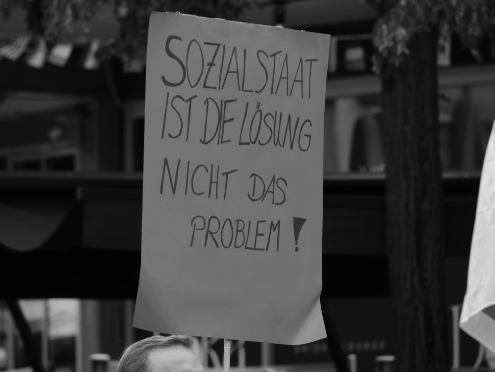 Das s/w-Foto zeigt ein einzelnes Plakat mit der Aufschrift "Sozialstaat ist die Lösung nicht das Problem!" im Hintergrund unscharf die Häuserfront.