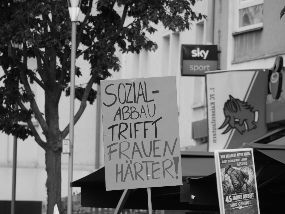 Das s/w-Foto zeigt zwei Demoplakate: "Sozialabbau trifft Frauen härter!" und "45 Jahre Arbeit sind Genug." Dahinter die Werbung einer Bar mit einer Fahne vom 1. FC Kaiserslautern.