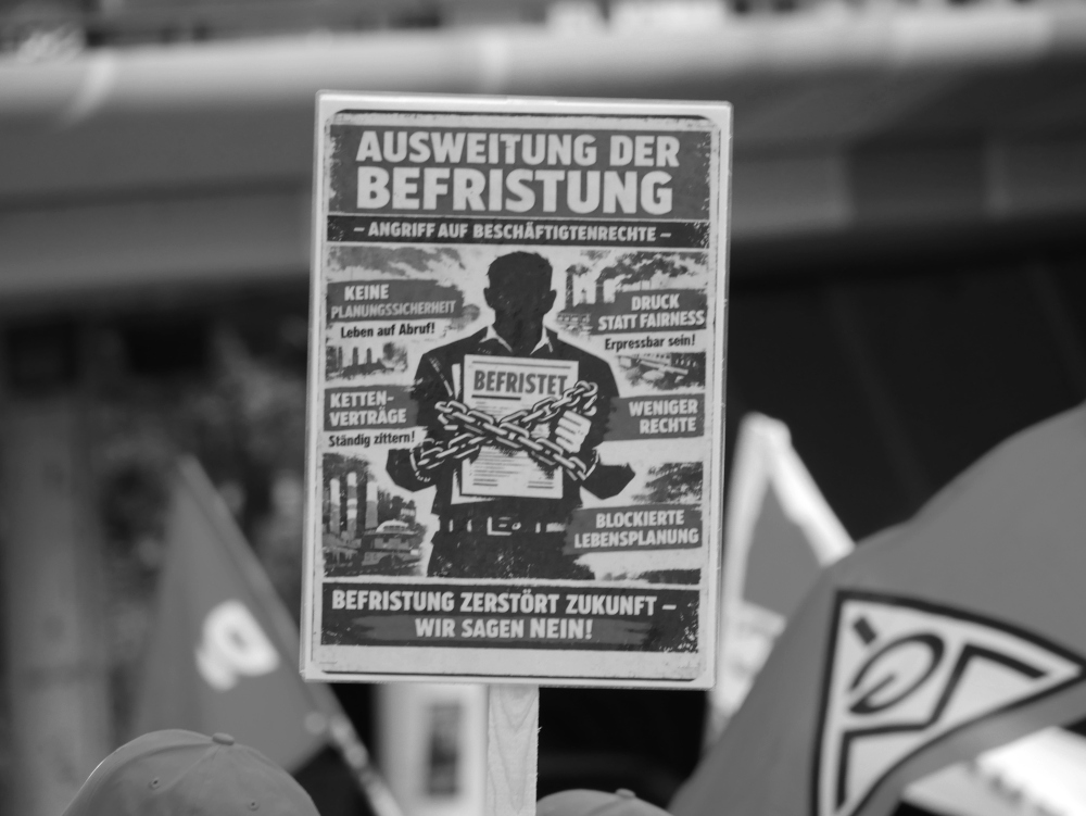 Das s/w-Foto zeigt ein Protestplakat keine eine Ausweitung der Befristung von Arbeitsverhältnissen zwischen Fahnen. Erkennbar ist die Fahne der IG Metall.